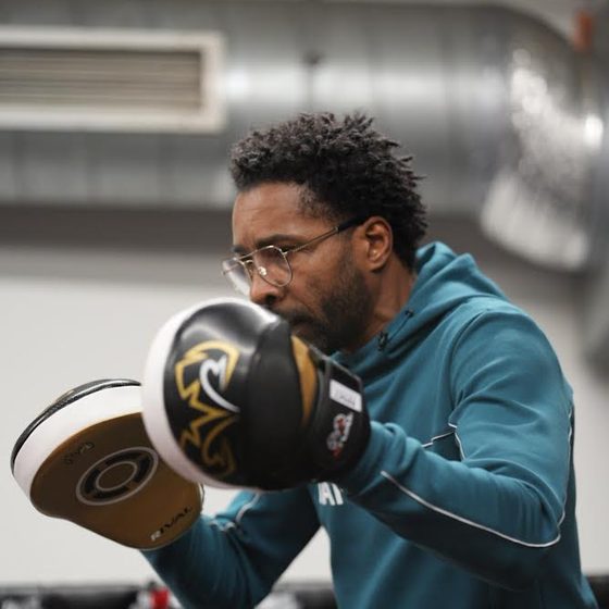 Een trainer met bril en blauwgroene hoodie houdt twee bokspads omhoog